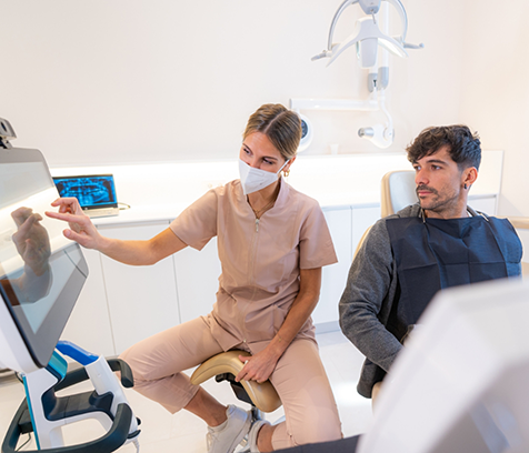 Dentist with mask sitting next to patient and pointing to monitor